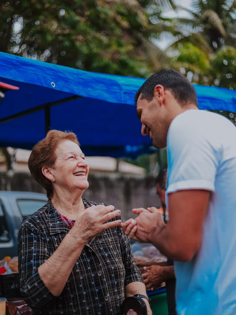 Serginho Vidigal segurando as mãos de uma senhora sorridente em uma feira livre
